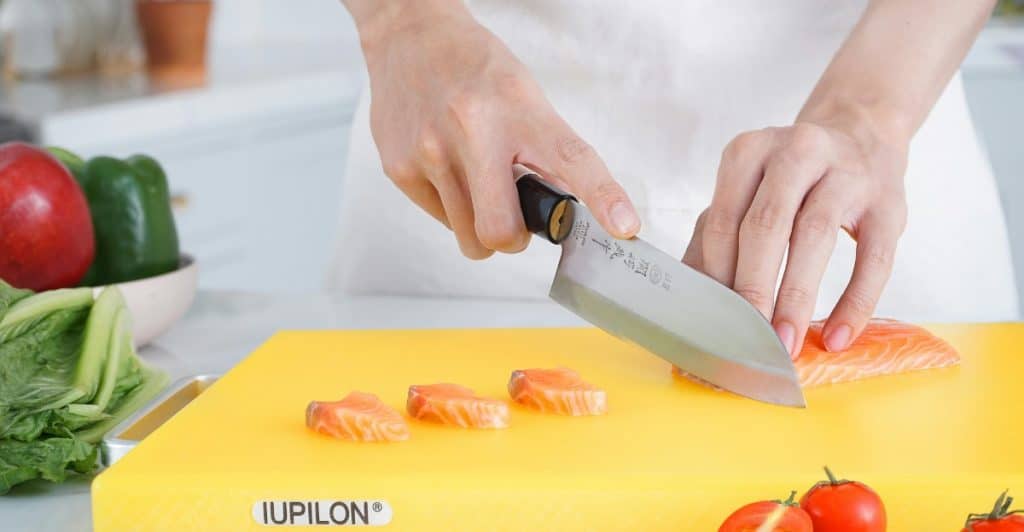 Close-up of hands holding a large Japanese-style knife, precisely slicing a piece of salmon on a yellow cutting board next to cherry tomatoes and other vegetables.
