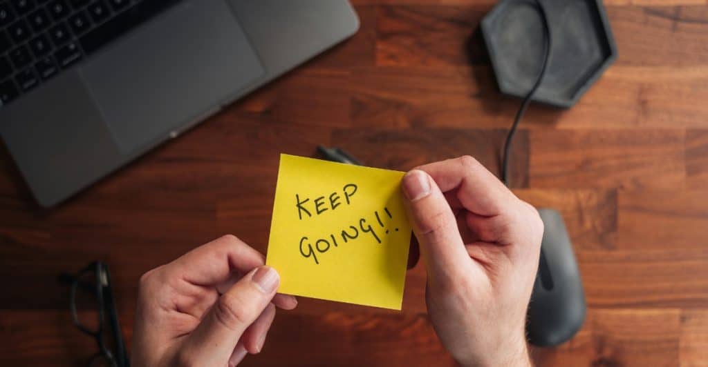 Hands holding a yellow sticky note with "KEEP GOING!!" written on it, with a laptop, mouse, and glasses on a wooden desk in the background.