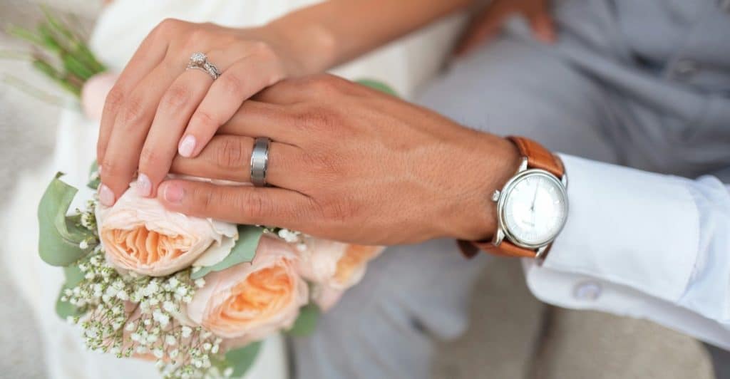 A close-up shot focuses on the hands of a married couple, with the woman's hand, adorned with an engagement ring and wedding band, resting gently on the man's hand, which wears a wedding band and a brown-strapped watch.