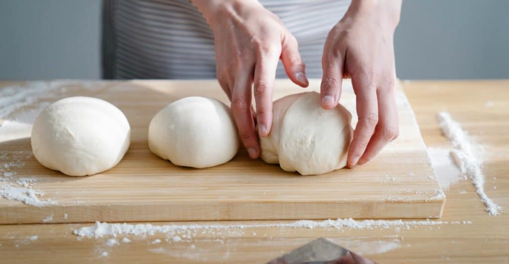 A person's hands wearing an apron, gently shaping three rounds of dough on a wooden cutting board dusted with flour, with a dough scraper nearby.