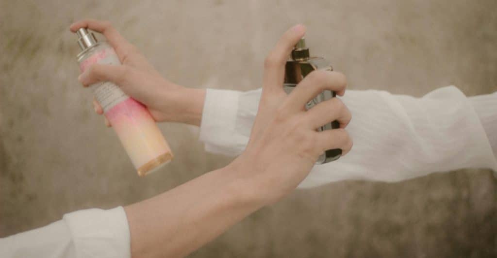 Two hands, one holding a pink and white spray bottle and the other holding a black spray bottle, both releasing mist.