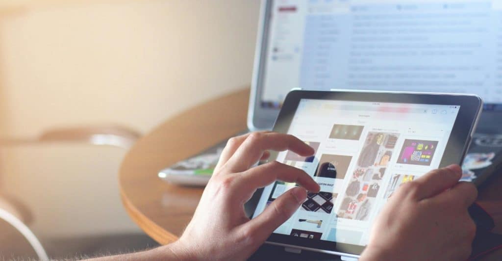 A person's hands touching a tablet screen, with a laptop in the background on a wooden table, in a brightly lit room.