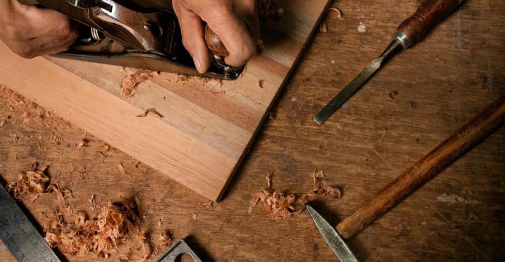 Hands using a hand plane on a piece of wood, with wood shavings, a chisel, a hammer, and a carpenter's square on a wooden surface.