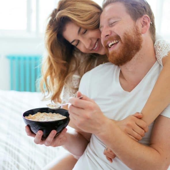A happy married couple embracing each other while the man eats from a bowl in bed.