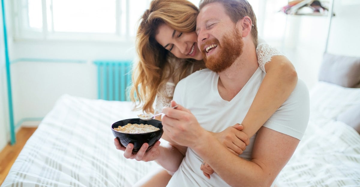 A happy married couple embracing each other while the man eats from a bowl in bed.