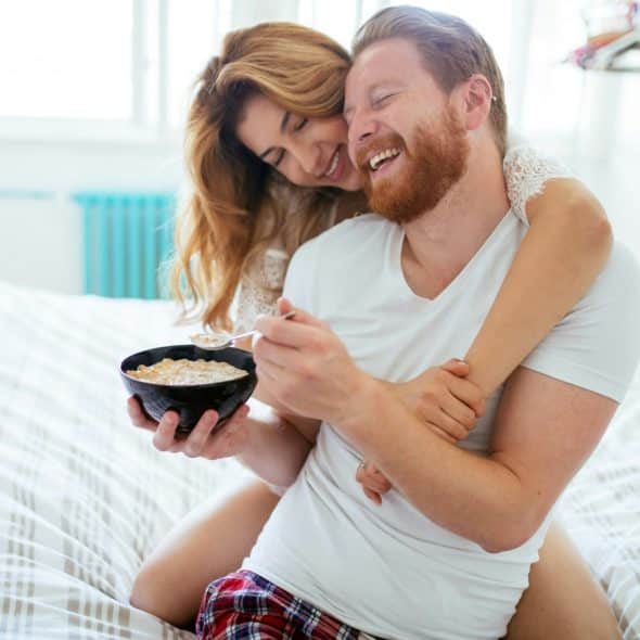 A happy married couple embracing each other while the man eats from a bowl in bed.