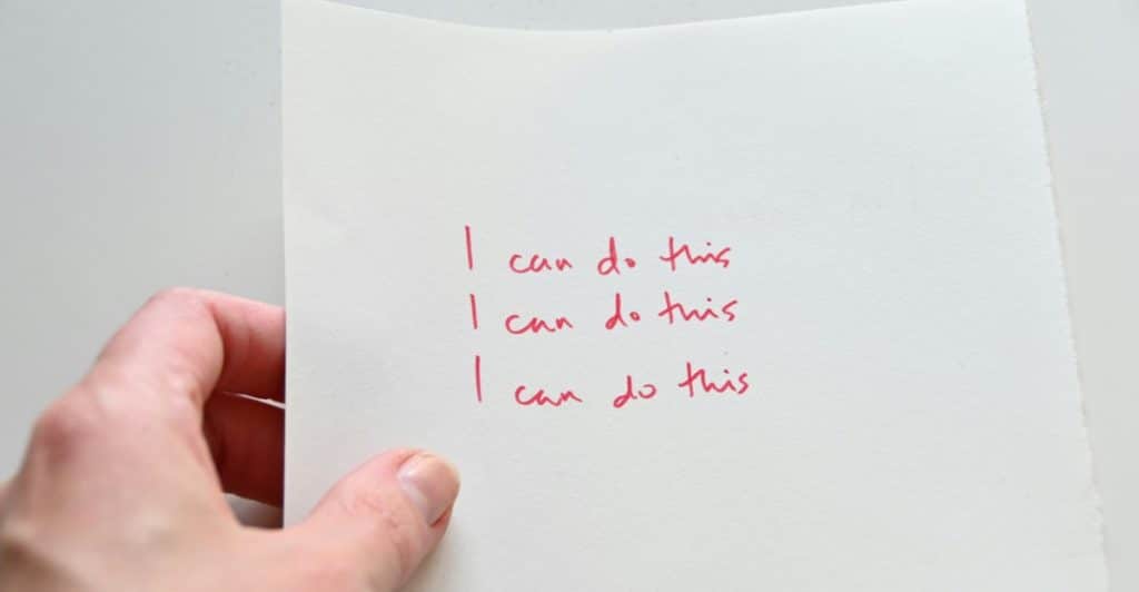 A hand holding a white paper with the words "I can do this" written three times in red ink.
