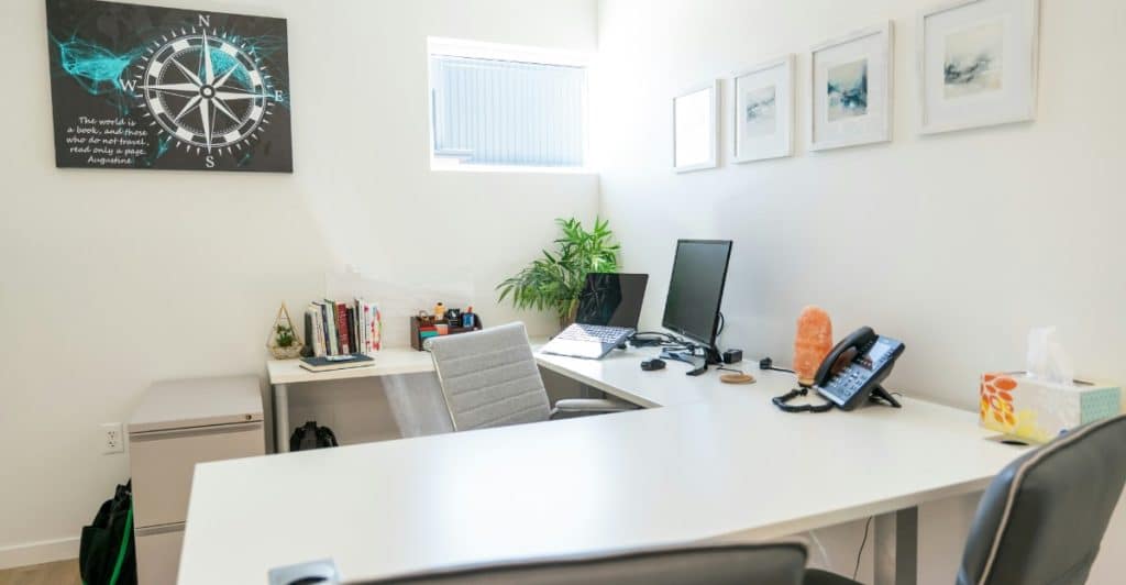 An L-shaped office desk with a computer, books, and a telephone, in a room with white walls and a compass art piece