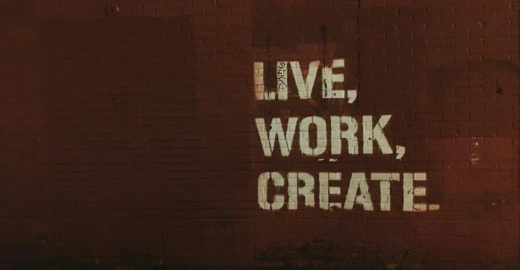 The words "LIVE, WORK, CREATE" spray-painted in white on a reddish-brown brick wall.