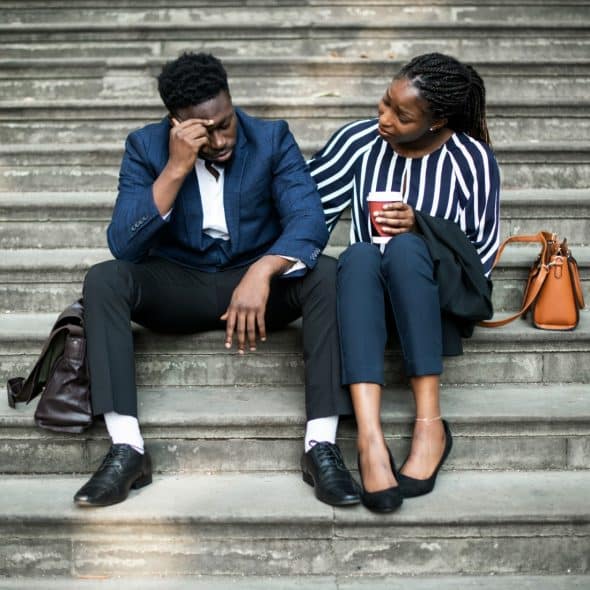 A woman sitting next to a distressed man on concrete steps, gently placing her hand on his shoulder while holding a cup.