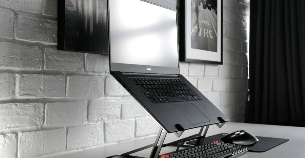 A black laptop elevated on a silver adjustable stand, with headphones and a keyboard on a white desk.