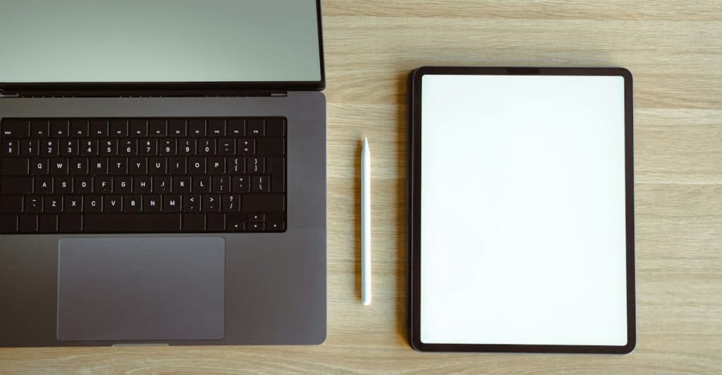 A laptop, a tablet with a blank screen, and a stylus are arranged on a wooden desk.