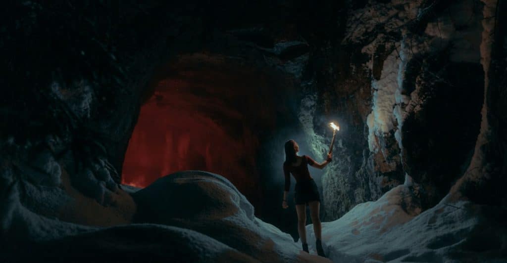 A person resembling Lara Croft standing in a snowy, dark cave, holding a torch that illuminates the surrounding ice and rock formations.