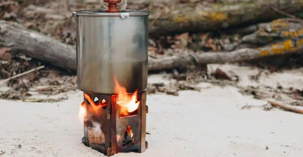 A lightweight camping stove burning beneath a metal pot, cooking outdoors on a sandy surface.