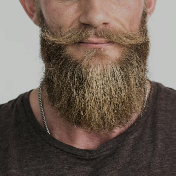 A close-up of a man with a long, well-groomed light brown beard and a distinct handlebar mustache, wearing a dark v-neck shirt.