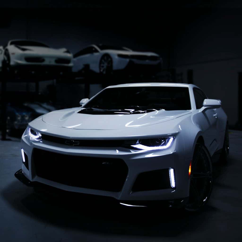 A white sports car with its lights on, parked in a dark garage with other luxury cars on lifts in the background.