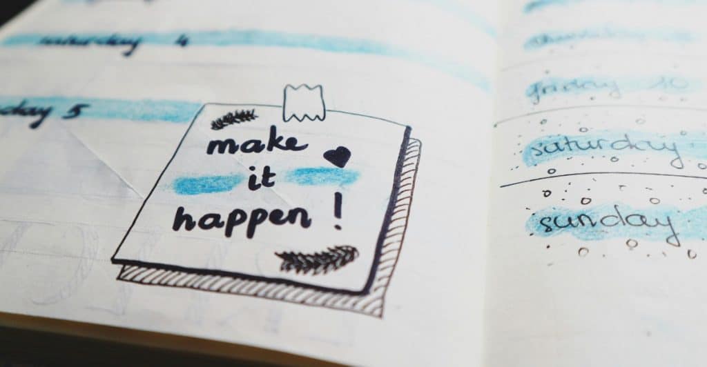 An open planner with a sticky note that says "make it happen!" and drawings of checkboxes and stars.