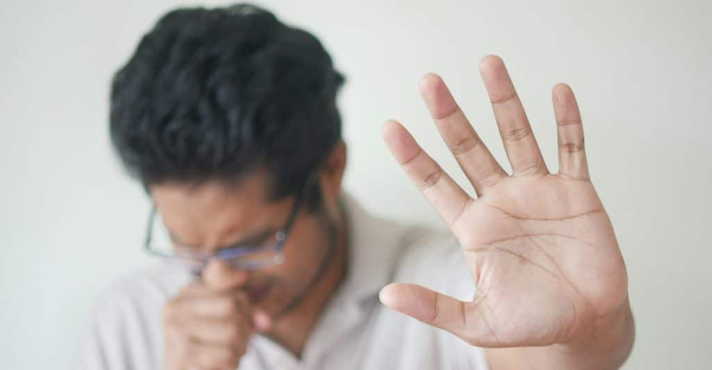 A Man coughing into his hand and holding his other hand up, palm out, to the screen.