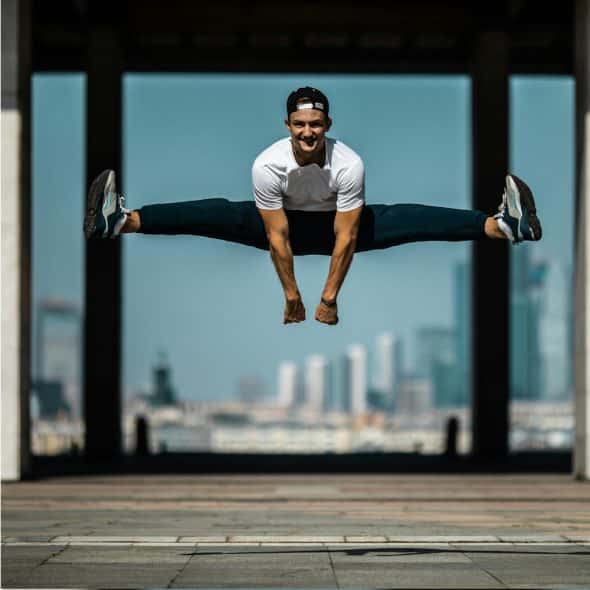 A man in athletic wear performing a split jump outdoors with a cityscape in the background.