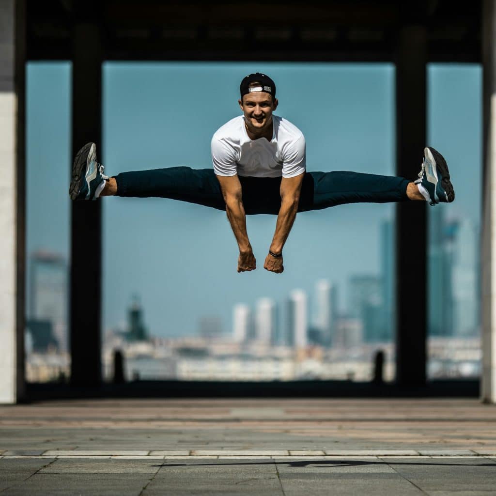 A man in athletic wear performing a split jump outdoors with a cityscape in the background.