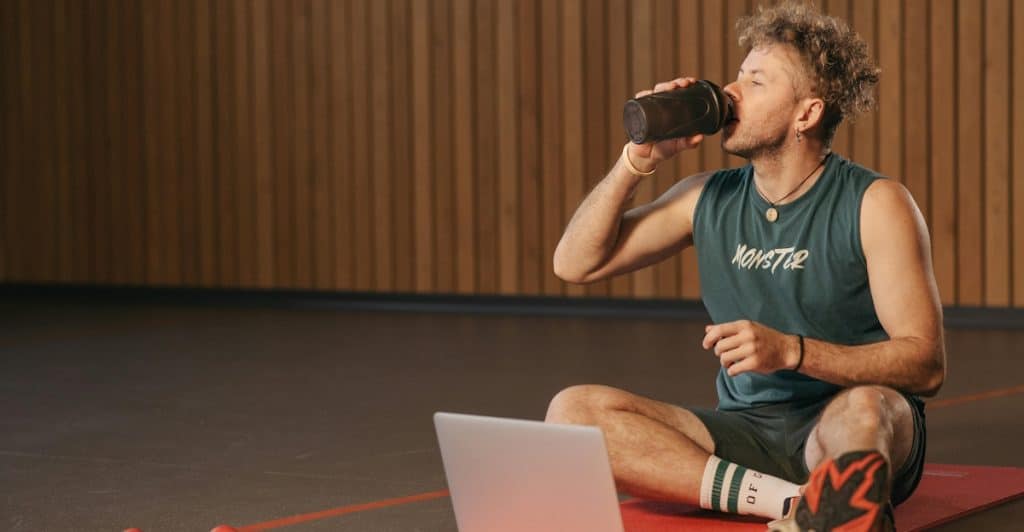 A man sitting on a yoga mat in a gym, drinking from a shaker bottle and looking at a laptop.