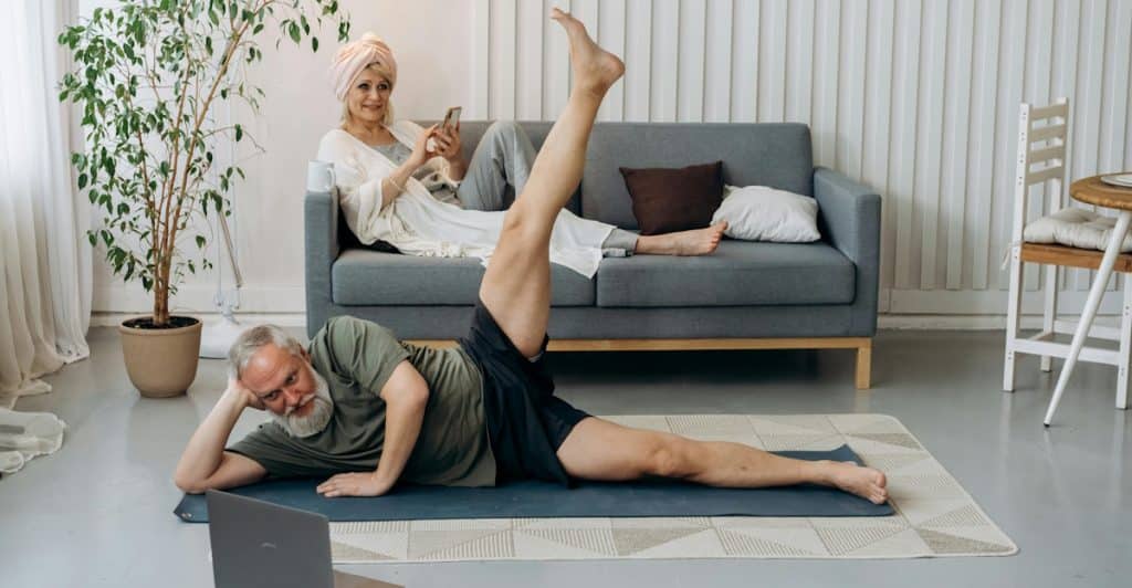 An elderly man lying on a yoga mat and looking at a laptop while exercising, and an elderly woman with a towel on her head sitting on a sofa Browse her phone.