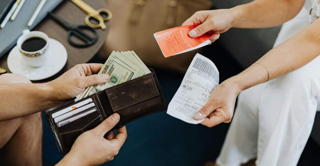 A close-up of a man's hands taking a stack of U.S. dollar bills from a brown leather wallet, while a woman's hands hold papers nearby on a table with a coffee cup.