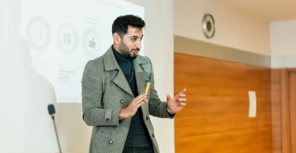 A man in a gray coat giving a presentation in front of a projected slide on business market segmentation.