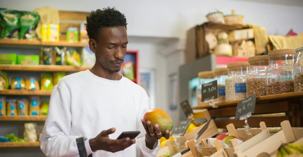 A man with a short beard holding a mango and looking at his phone in a grocery store aisle filled with produce.