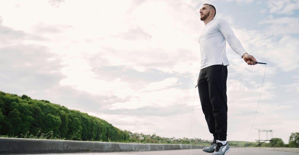 A man jumping rope on a paved surface with a green landscape and cloudy sky in the background.