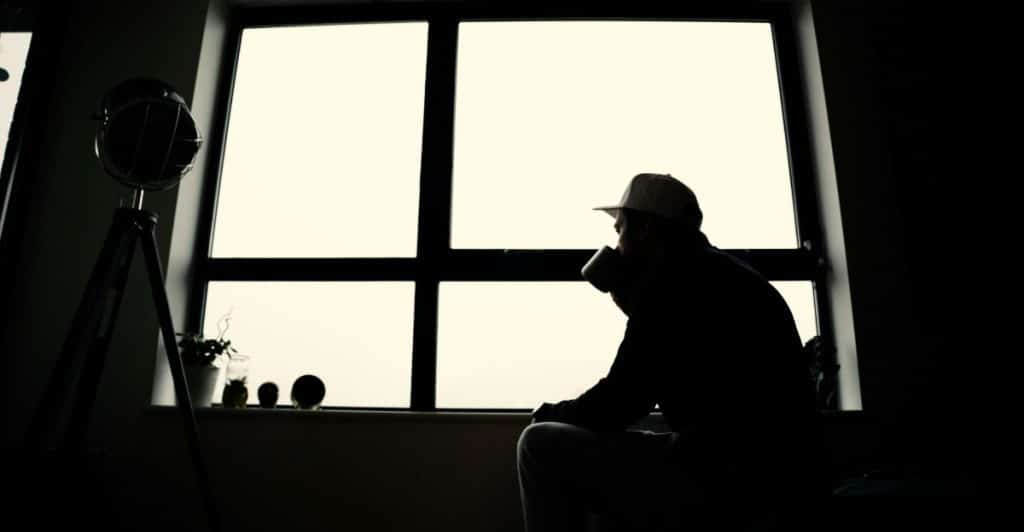 A man in a cap and dark clothing is silhouetted against a large window, holding a cup and looking out, creating a contemplative scene.