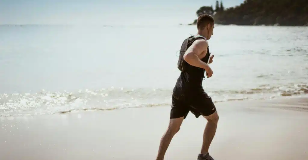 A man in a black tank top and black shorts running on a beach next to the ocean.