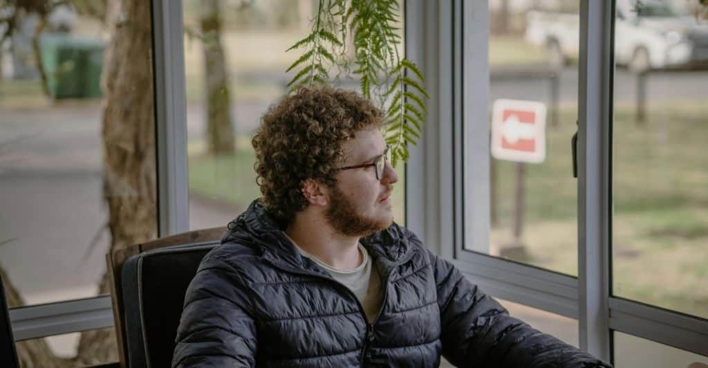 A man with curly hair and glasses sitting indoors, looking out of a window.