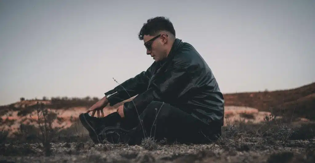 A man in a leather jacket sitting on rocky ground at dusk.