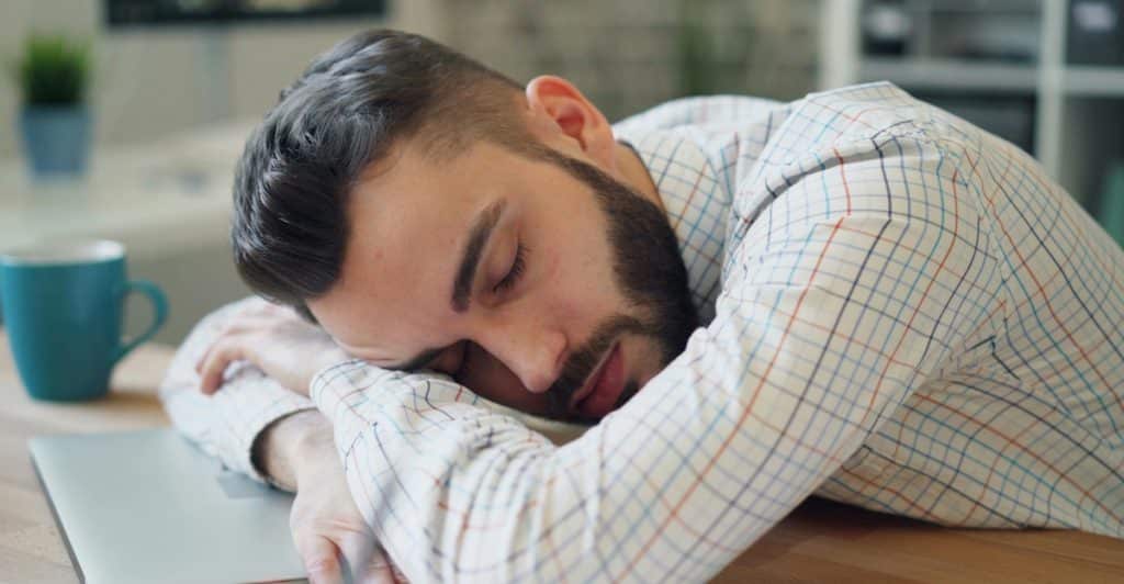 A man in a checkered shirt sleeping with his head resting on his arms at a desk.