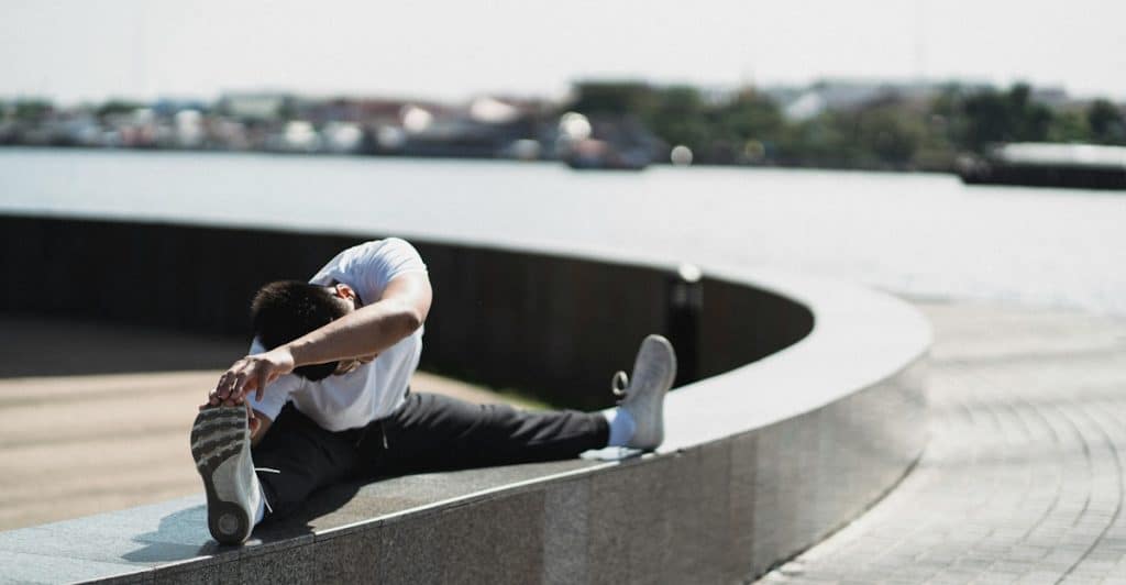 A man stretching his leg while sitting on a curved outdoor structure with a body of water in the background.
