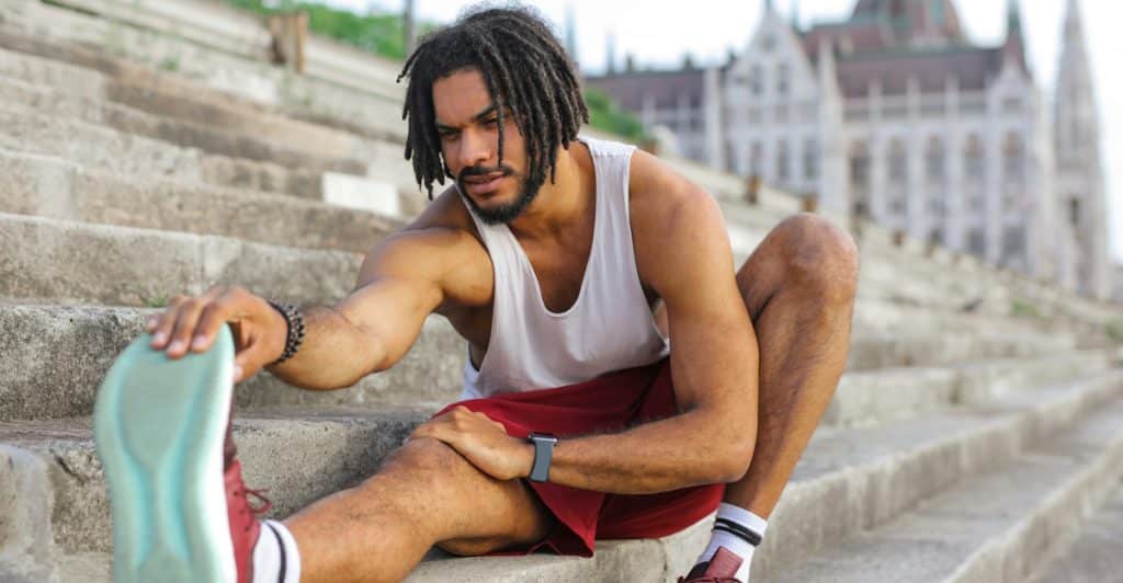 A man with dreadlocks stretching his leg while sitting on outdoor steps.