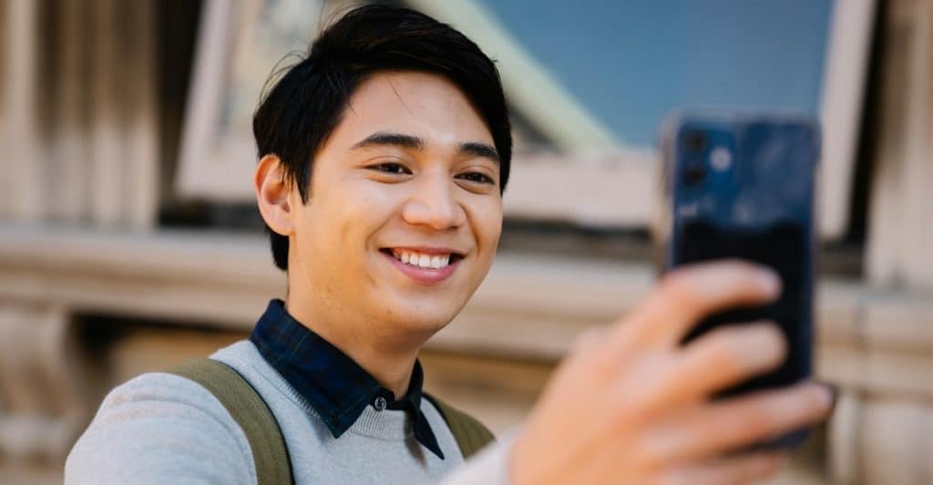 A young man with dark hair smiling and taking a selfie outdoors with a blue sky in the background.