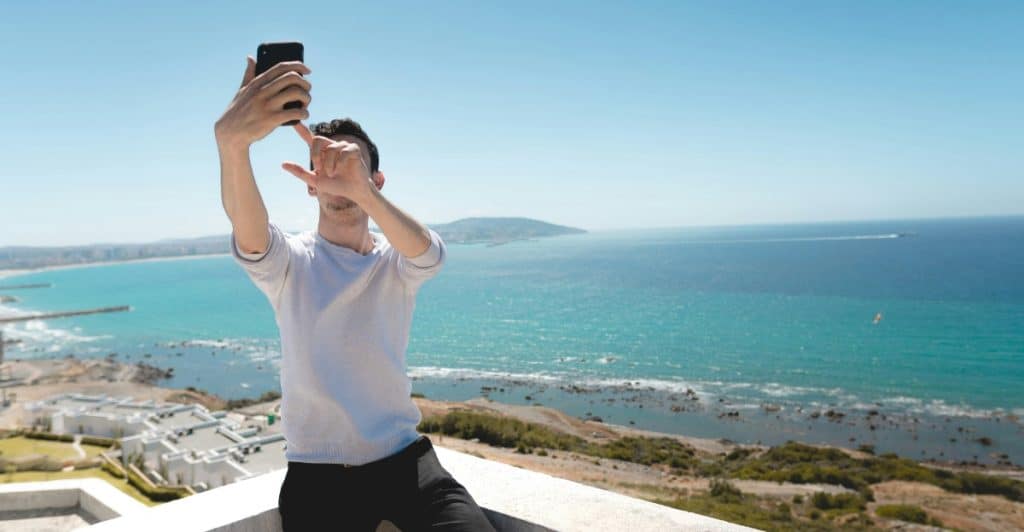A man taking a selfie with a scenic view of the ocean and a town in the background.