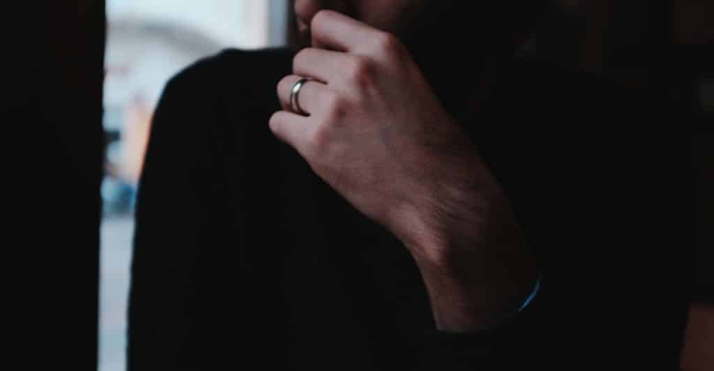A close-up of a man's left hand, adorned with a wedding ring, raised to his chin as he stands in a dimly lit room, with his face obscured.