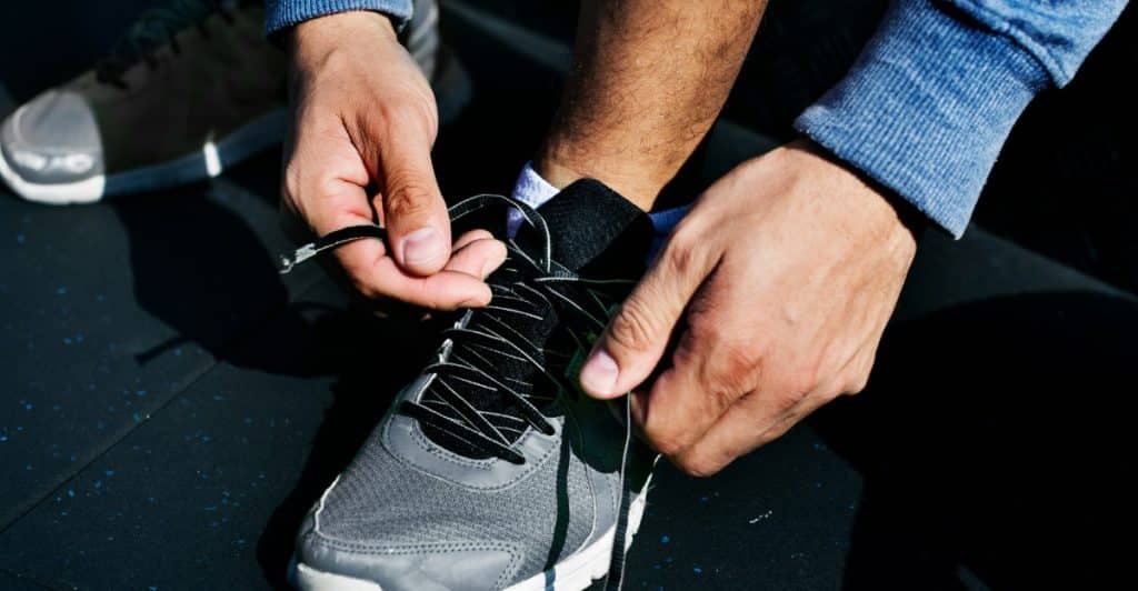 A man's hands tying the black laces of a grey sneaker, with his feet on a dark textured surface.