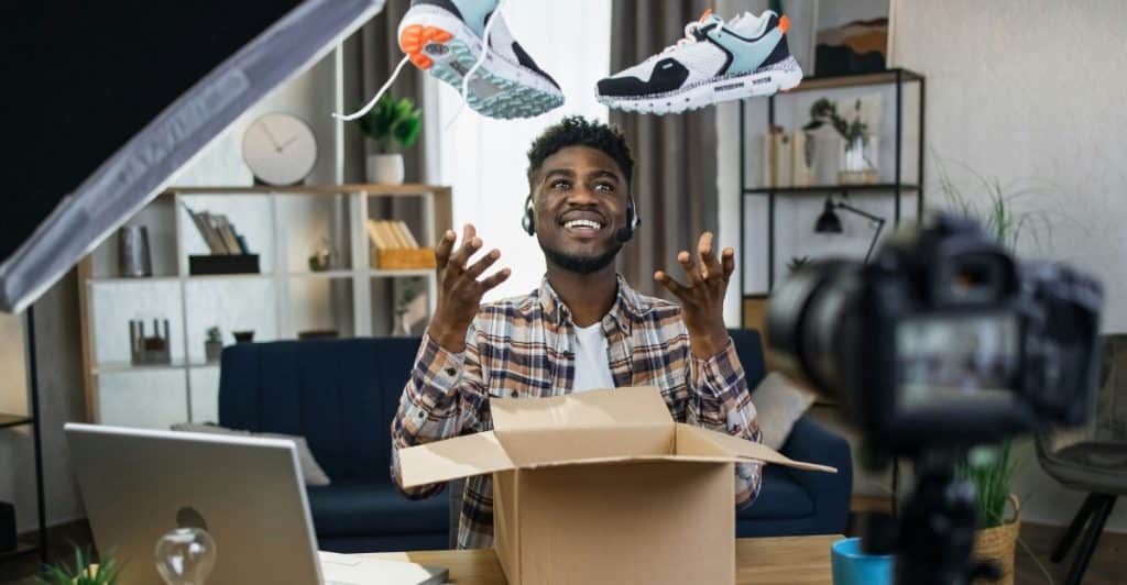 A man with short dark hair and a beard, wearing a plaid shirt and headphones, sitting at a desk with a laptop and camera, happily unboxing sneakers that are floating above him.