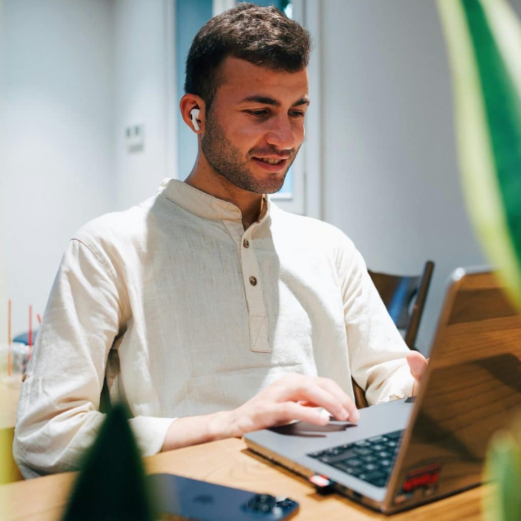 A man with short brown hair and a light-colored shirt is smiling while using a laptop at a table with plants nearby.