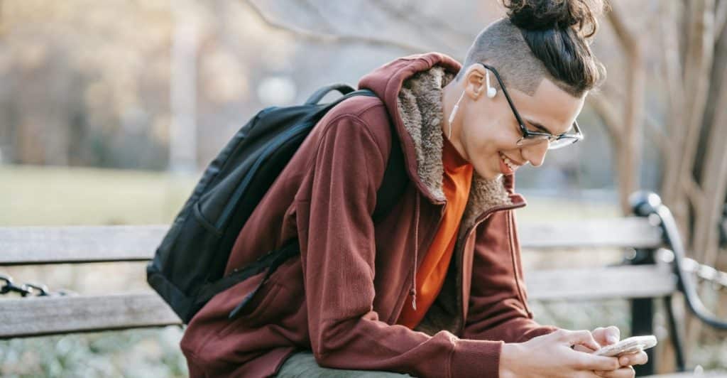 A man with a unique hairstyle sitting on a park bench and using his smartphone.
