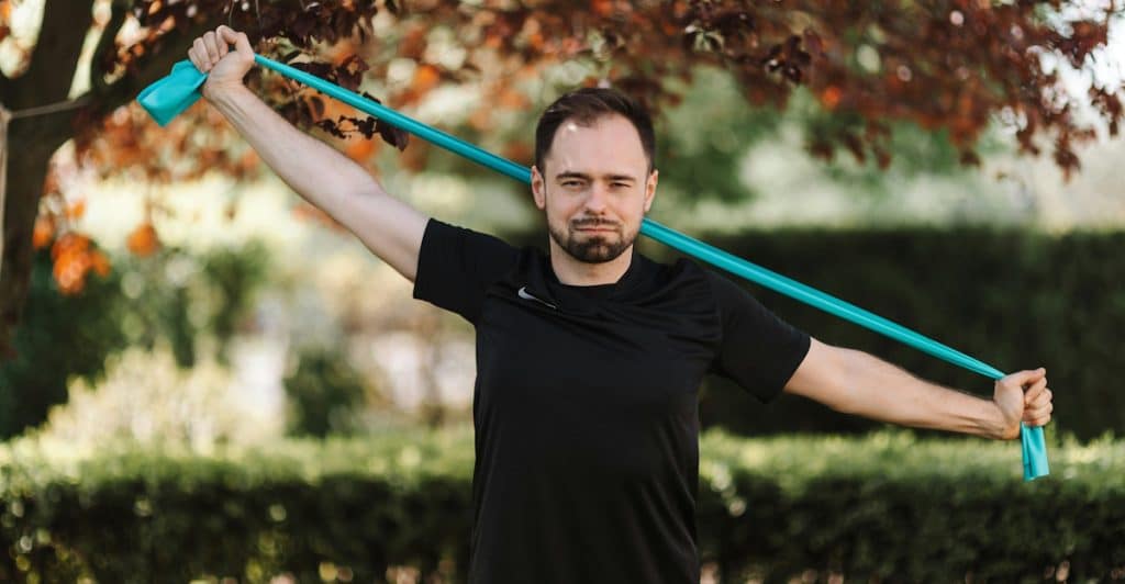 A man standing outdoors holding a blue resistance band with trees in the background.