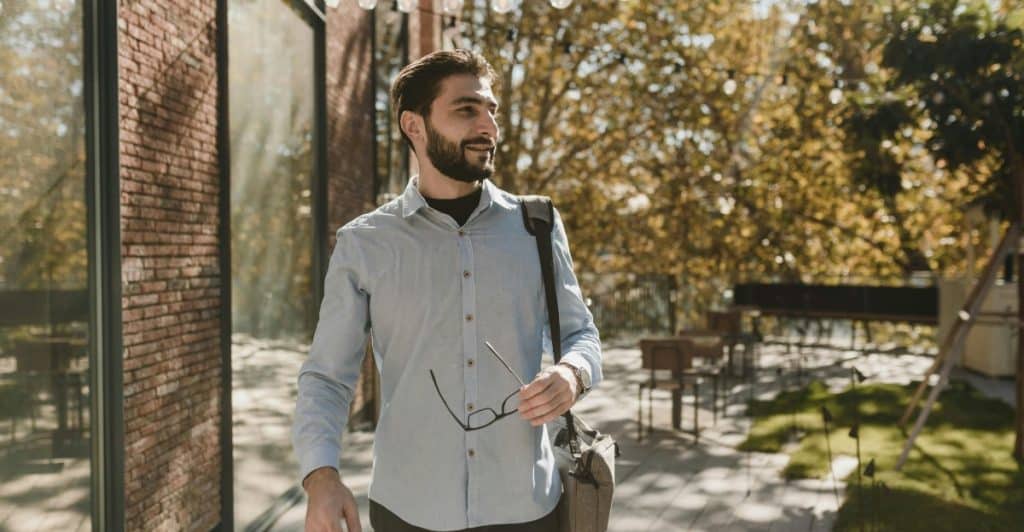 A man with a beard walking outdoors with a shoulder bag and holding glasses.