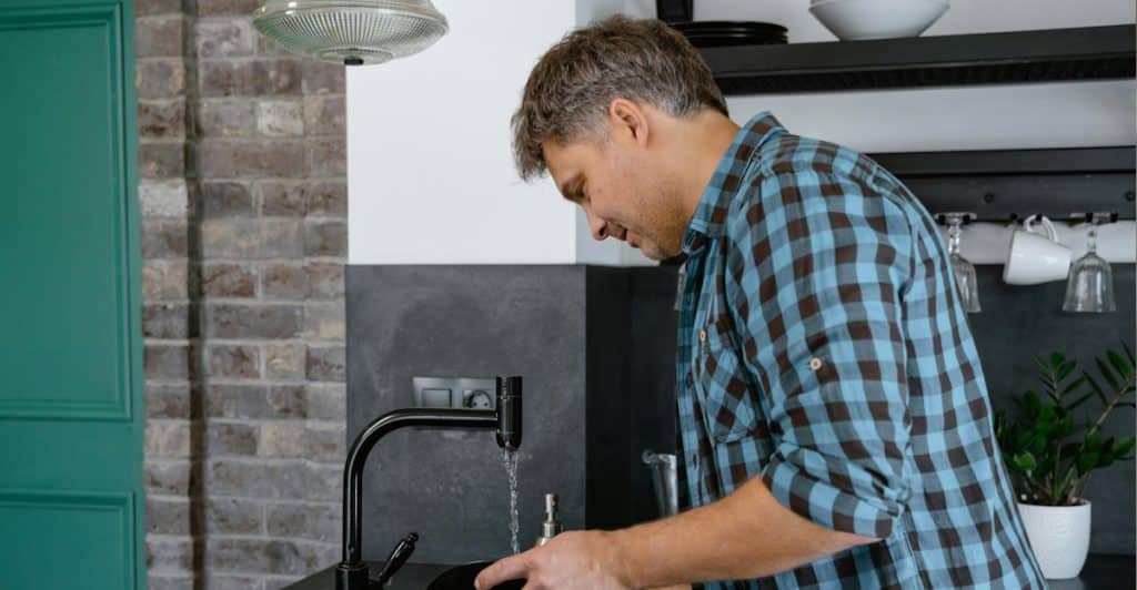 A man in a blue plaid shirt standing at a kitchen sink, washing a dark plate under running water.