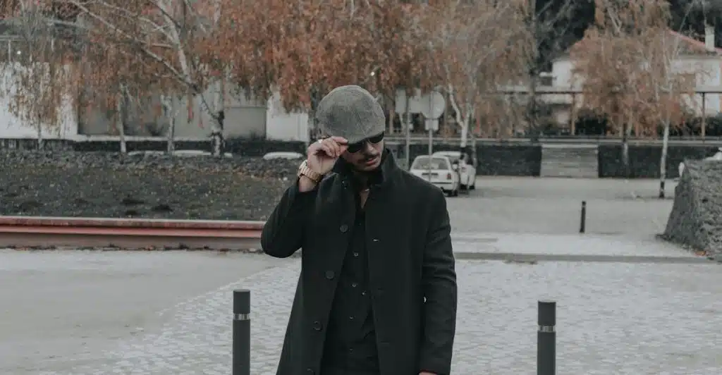 A stylish man in a dark coat and flat cap adjusting his sunglasses while walking through a cobblestone area.
