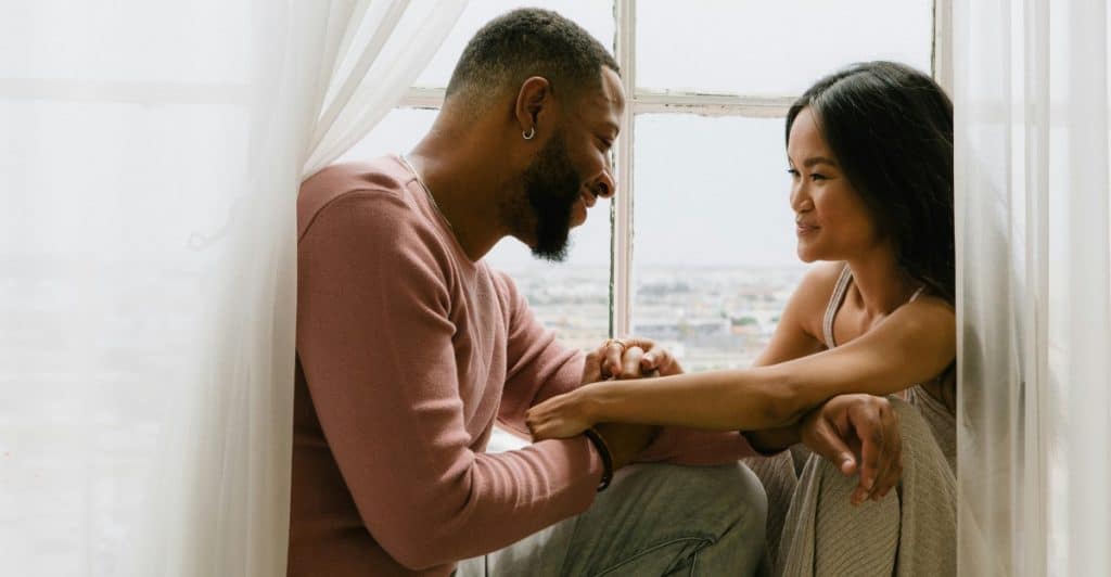 A man and a woman sitting by a window, holding hands and looking at each other, with natural light streaming in.