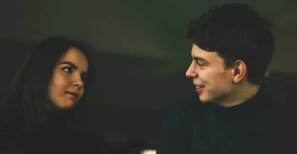 A man and a woman, both dressed in dark long-sleeved shirts, are engaged in conversation, looking at each other with soft expressions in a dimly lit indoor setting.